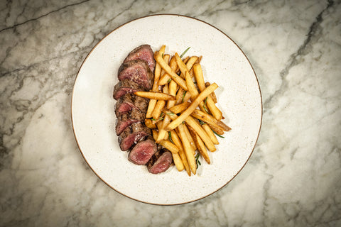 A plate with sliced venison striploin steak with chips on a marble countertop.