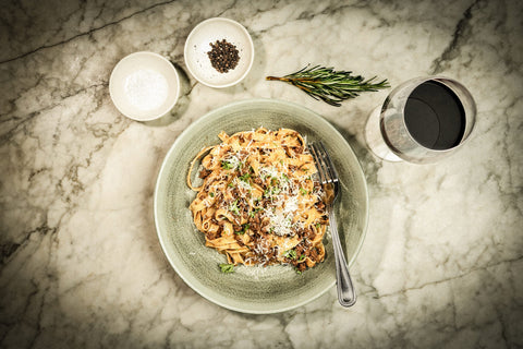 A bowl of pappardelle with venison ragu sprinkled with parmesan on a marble table with a glass of red wine.